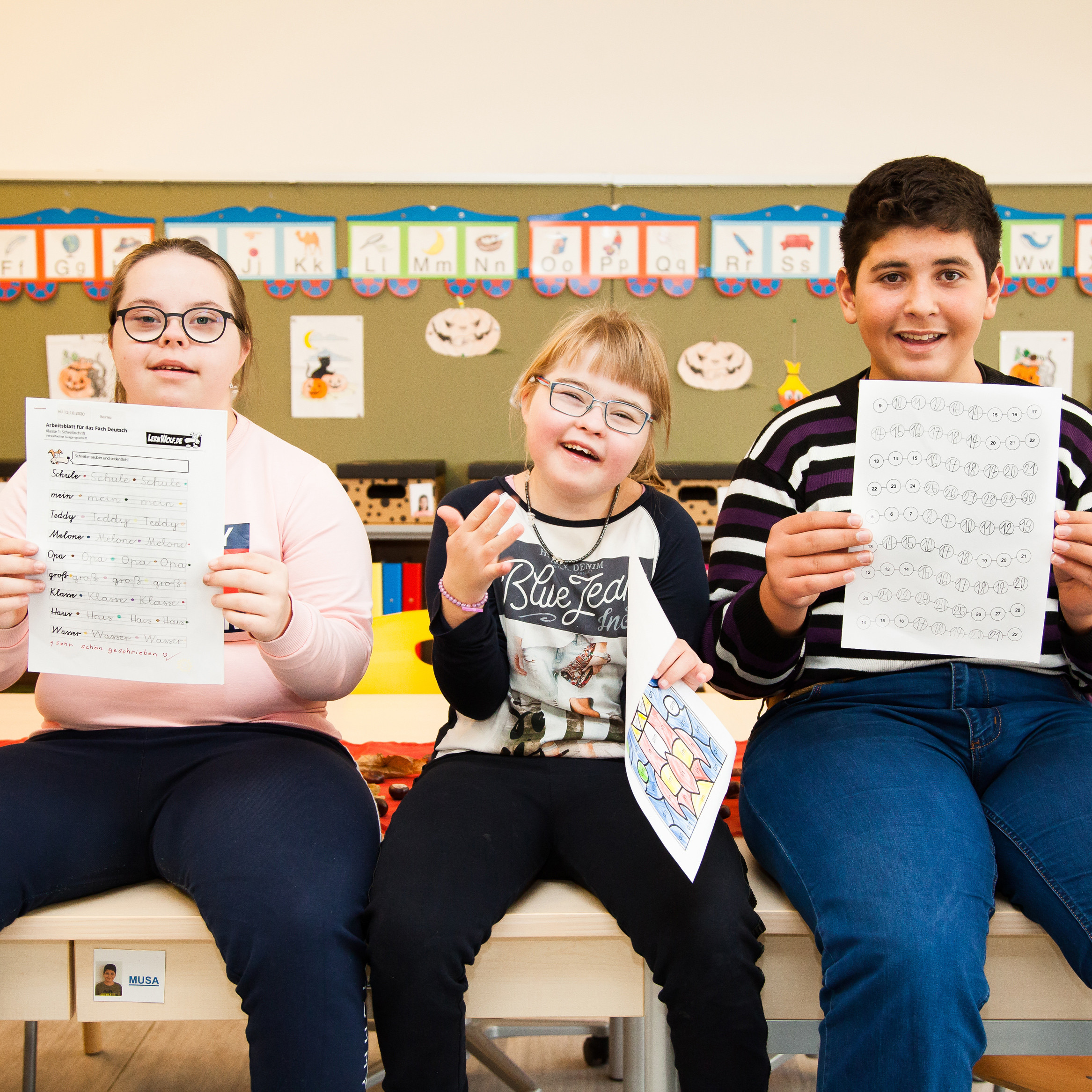 Drei Kinder mit und ohne Behinderungen lachen in die Kamera. Sie sitzen auf Tischen und halten Übungsblätter hoch.