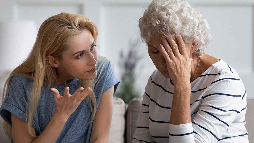 Eine jüngere Frau im Gespräch mit älterer Dame, die sich überfordert das Gesicht hält.