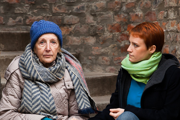 Mitarbeiterin der Diakonie setzt sich zu einer älteren Frau, die in der Kälte auf einer Stiege sitzt