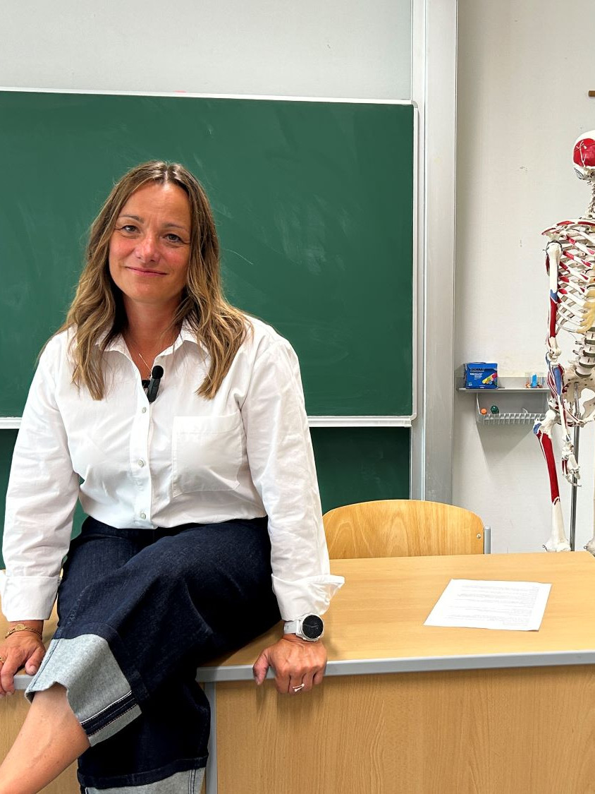 Frau sitzt auf Lehrerpult. Im Hintergrund grüne Schultafel und Skelett.