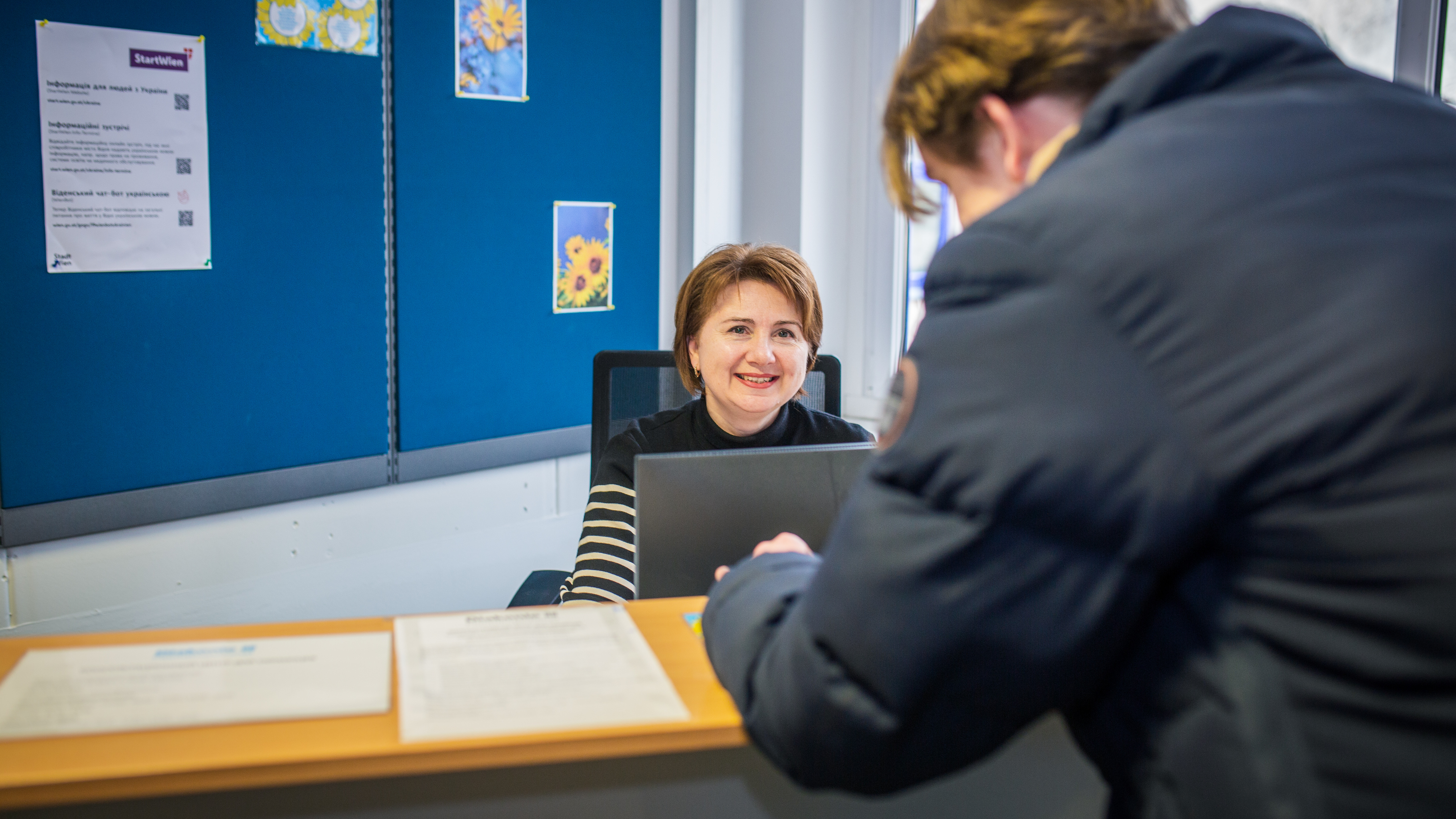 Eine lächelnde Frau sitzt hinter einem Empfangstresen am PC. Man sieht eine weitere Person von hinten am Tresen stehen.