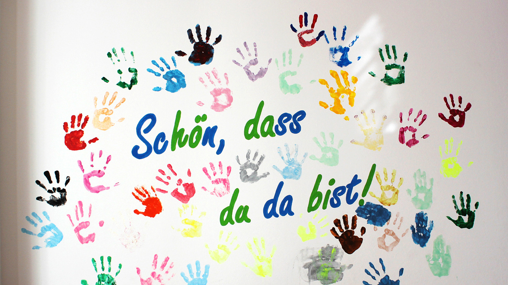 Viele bunte Farabdrücke von Kinderhände auf einer weißer Wand, dazu folgender Schriftzug: Schön, dass du da bist!