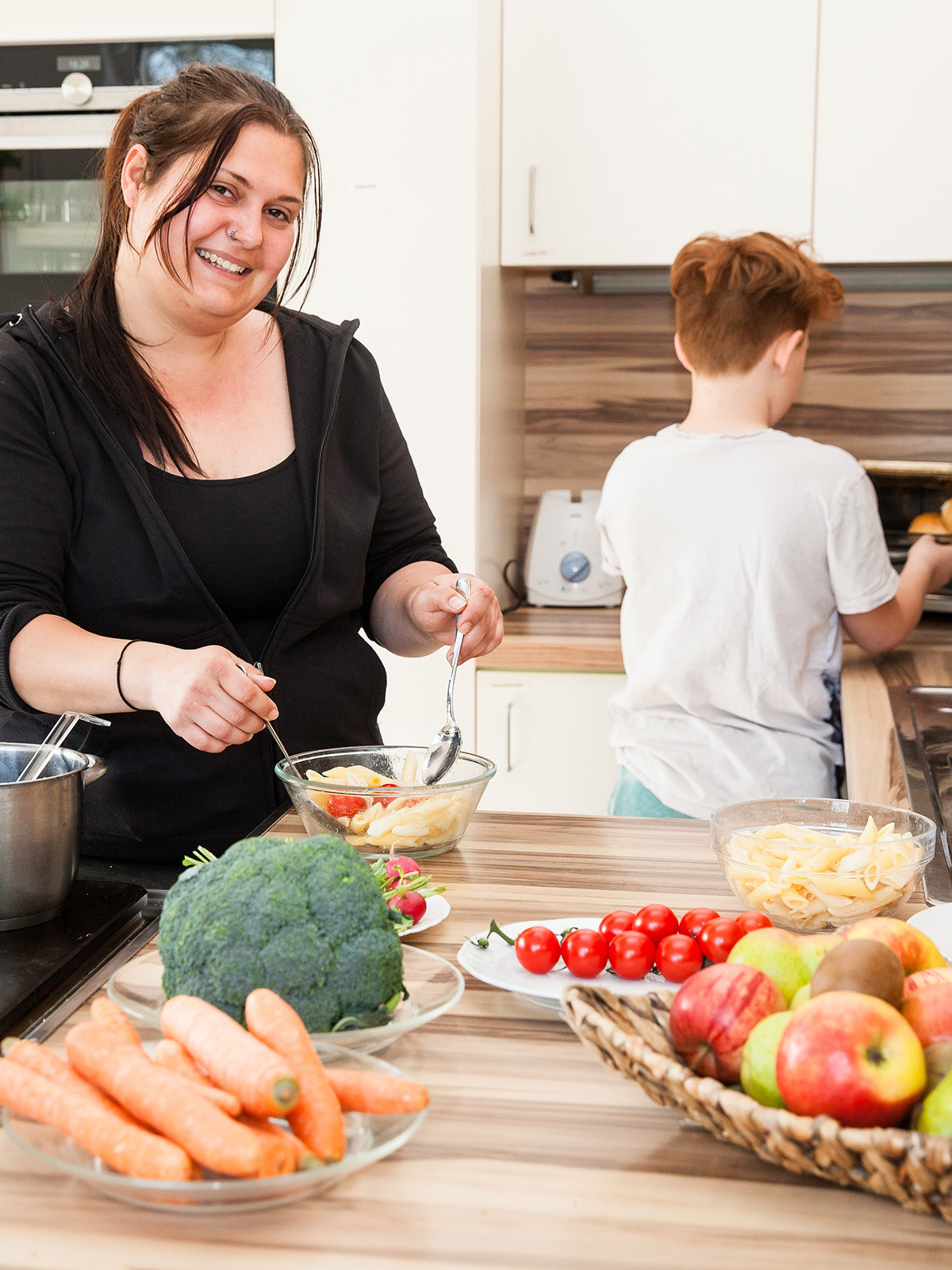 Pädagogin beim Zubereiten einer Mahlzeit in der Küche. Am Küchenblock stehen Obst, Gemüse, Küchenkräuter und Nudeln. Ein Junge schiebt Semmeln in einen Backofen.