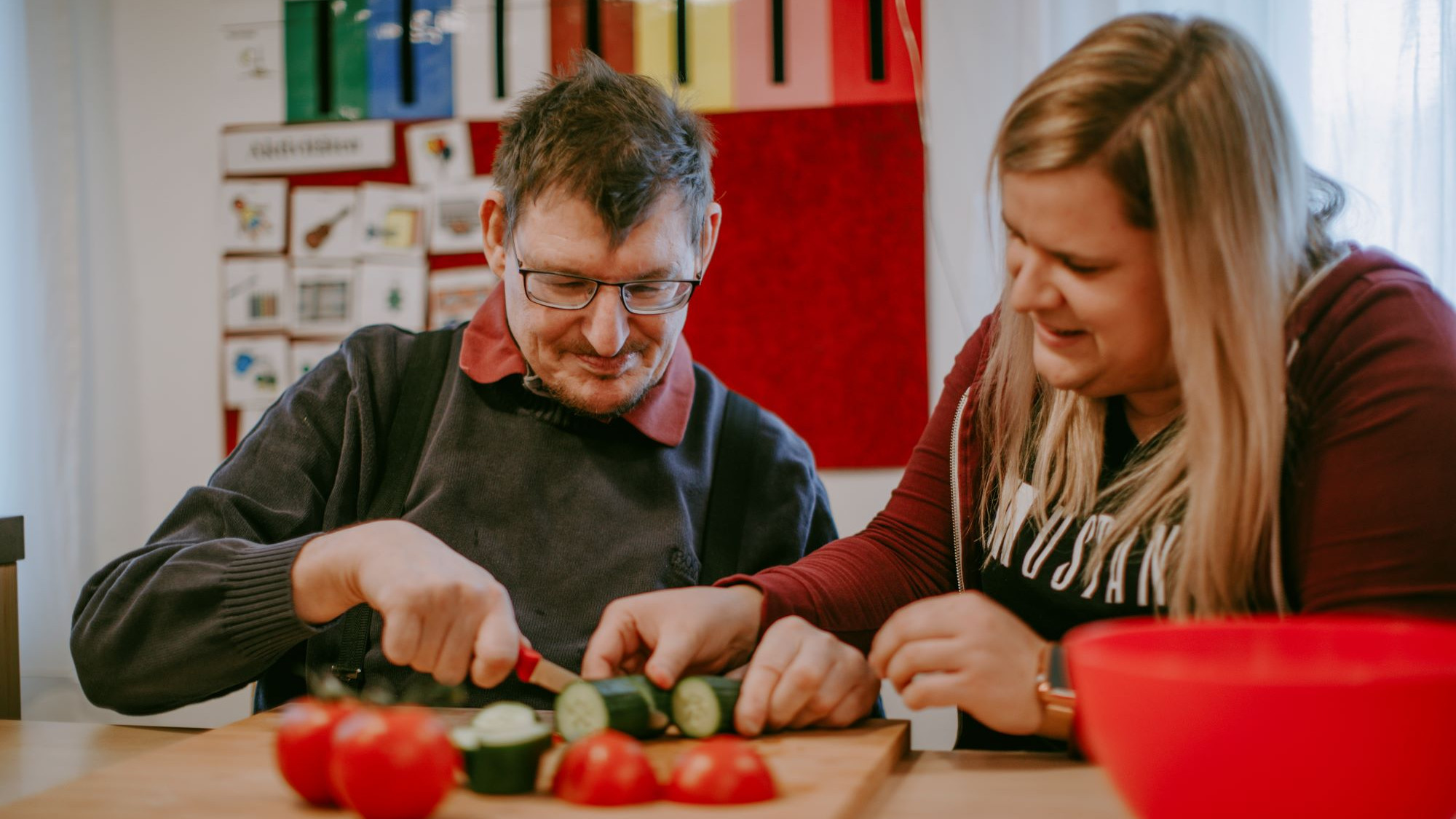 Zwei Personen sitzen bei Tisch, eine Person schneidet Gurken und Tomaten auf