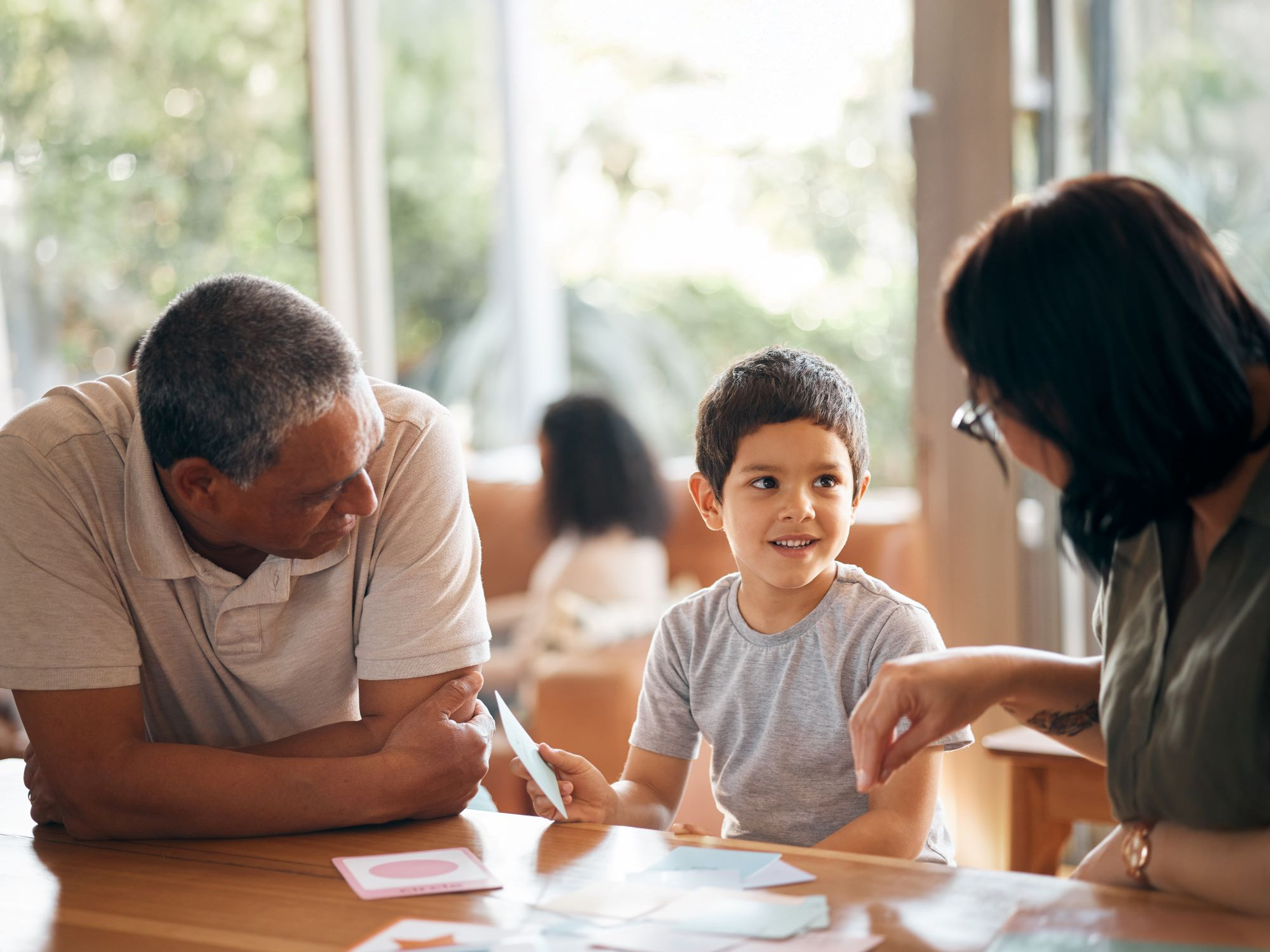 Kleiner Junge sitzt mit zwei Erwachsenen am Tisch und zeigt Kärtchen