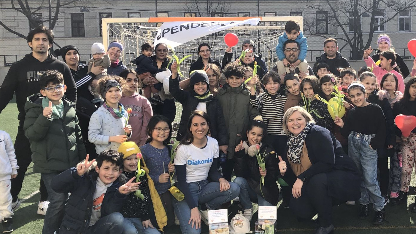 Volksschulkinder mit Eltern und Lehrer:innen auf einem Sportplatz vor der Schule. Einige halten rote herzförmige Luftballons
