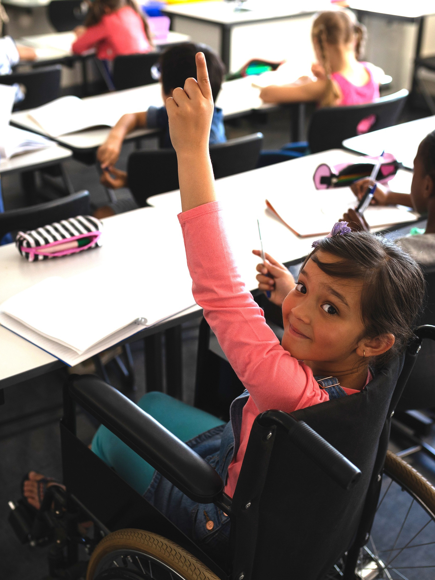 Eine Klasse mit jungen Schüler:innen. Ein Mädchen im Rollstuhl zeigt auf und blickt verschmitzt in die Kamera.