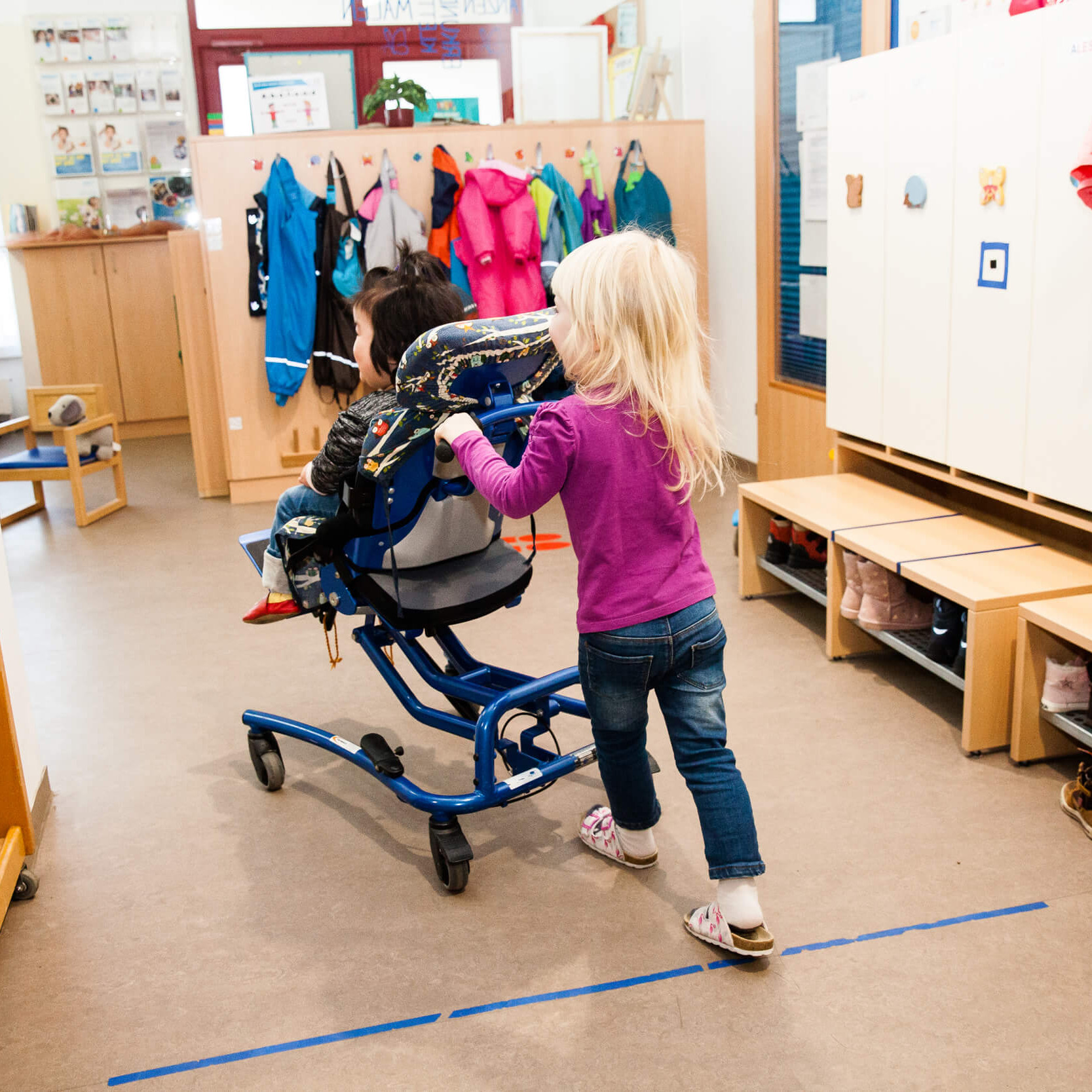 Ein Mädchen schiebt ein Mädchen im Rollstuhl durch einen Kindergarten.
