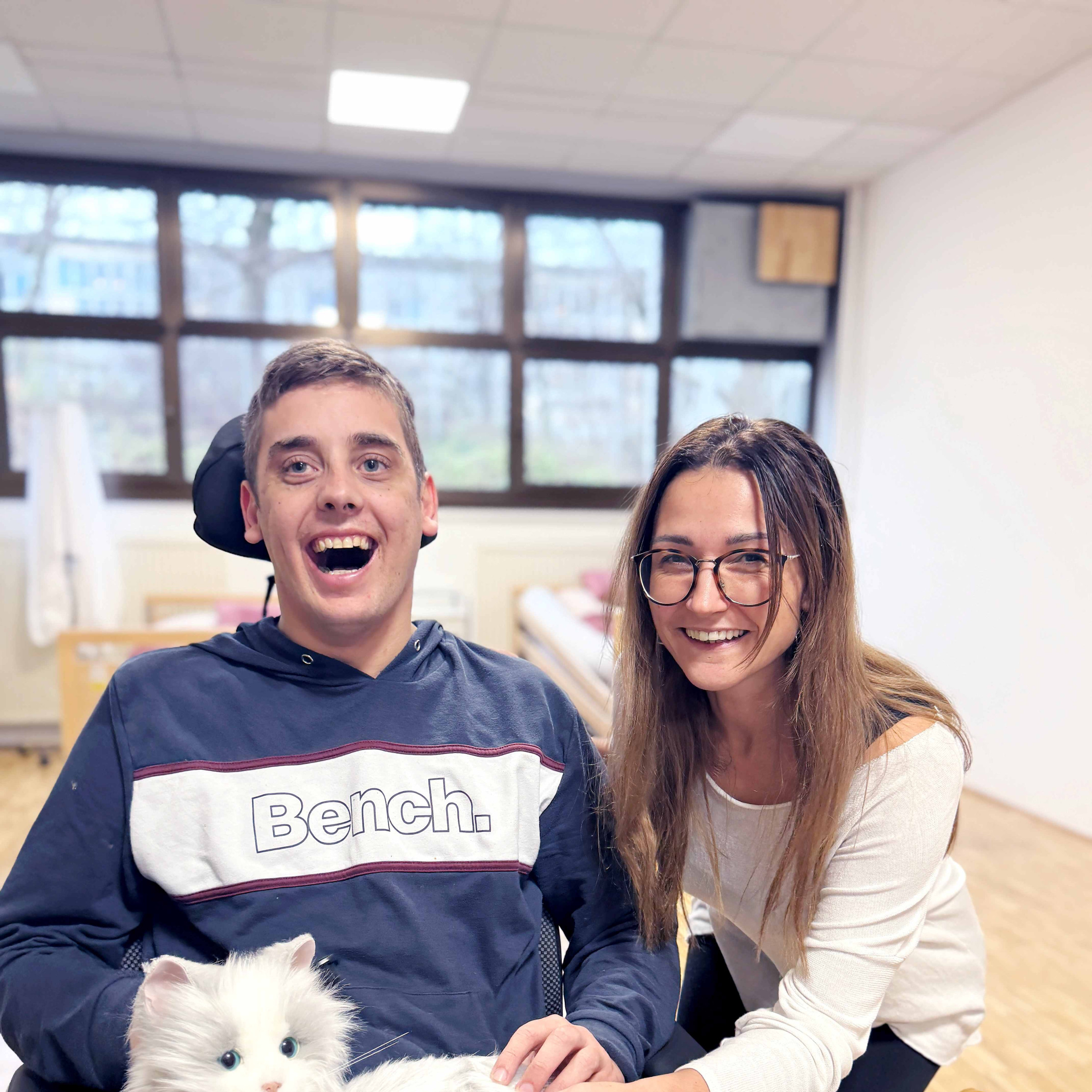 Zwei Personen aus dem inklusiven Lernzentrum lachen in die Kamera. Eine Person sitzt im Rollstuhl und hat eine Stofftierkatze am Schoß.