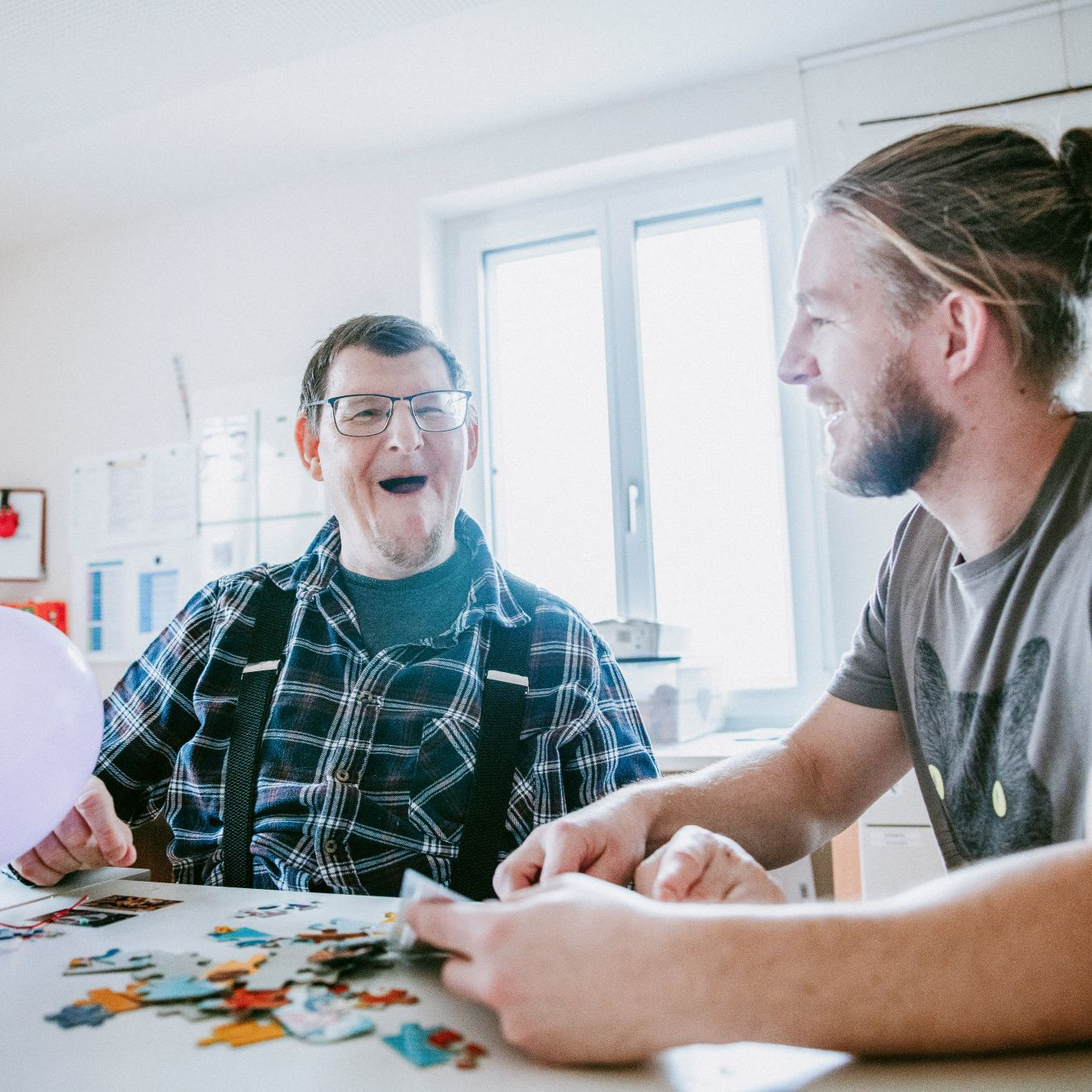 Zwei Personen sitzen nebeneinander bei Tisch, vor ihnen zwei Luftballons und Puzzlesteine