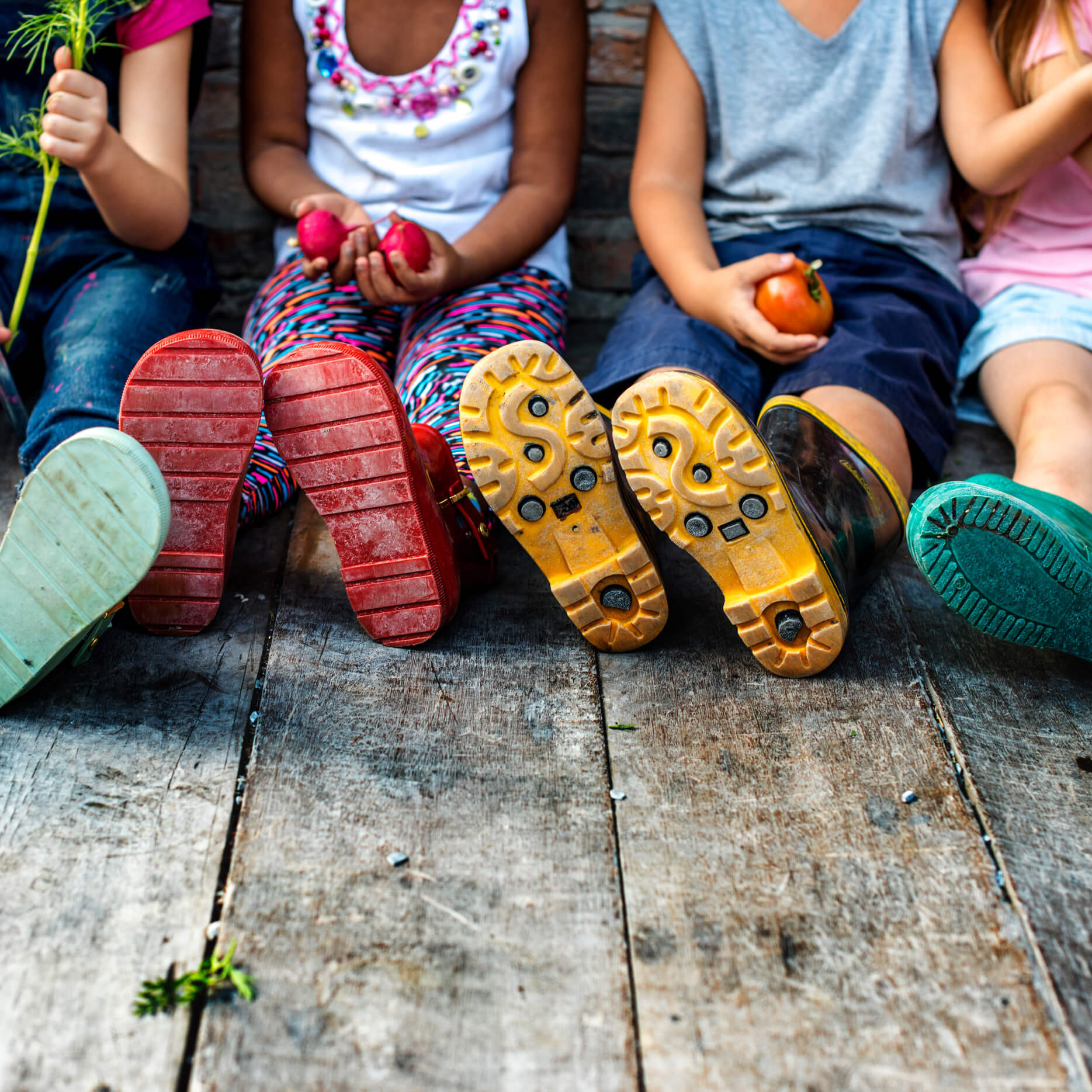 Mehrere Kinder mit bunten Gummistiefeln siitzen nebeneinander auf einem Weg aus Holzlatten und halten verschiedenes Gemüse in den Händen.