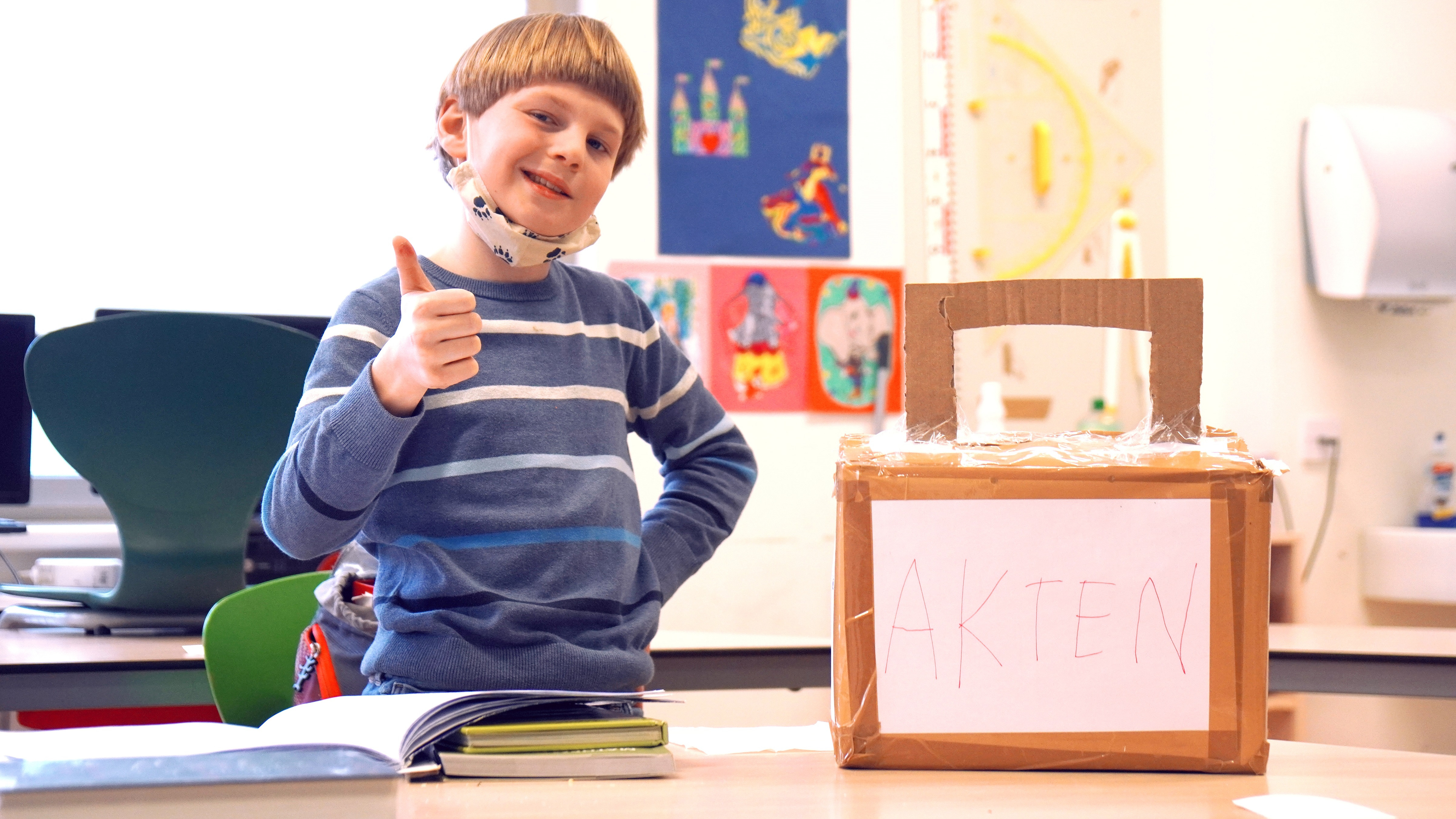 Ein junger Schüler zeigt mit dem Daumen nach oben und schaut stolz in die Kamera, neben ihm steht auf dem Schultisch eine selbstgebastelte Aktentasche aus Karton, auf der in großen Buchstaben Akten steht.
