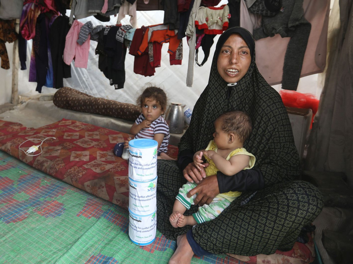 Frau sitzt mit zwei Kindern in Notzelt, mit drei Packungen Babymilchpulver