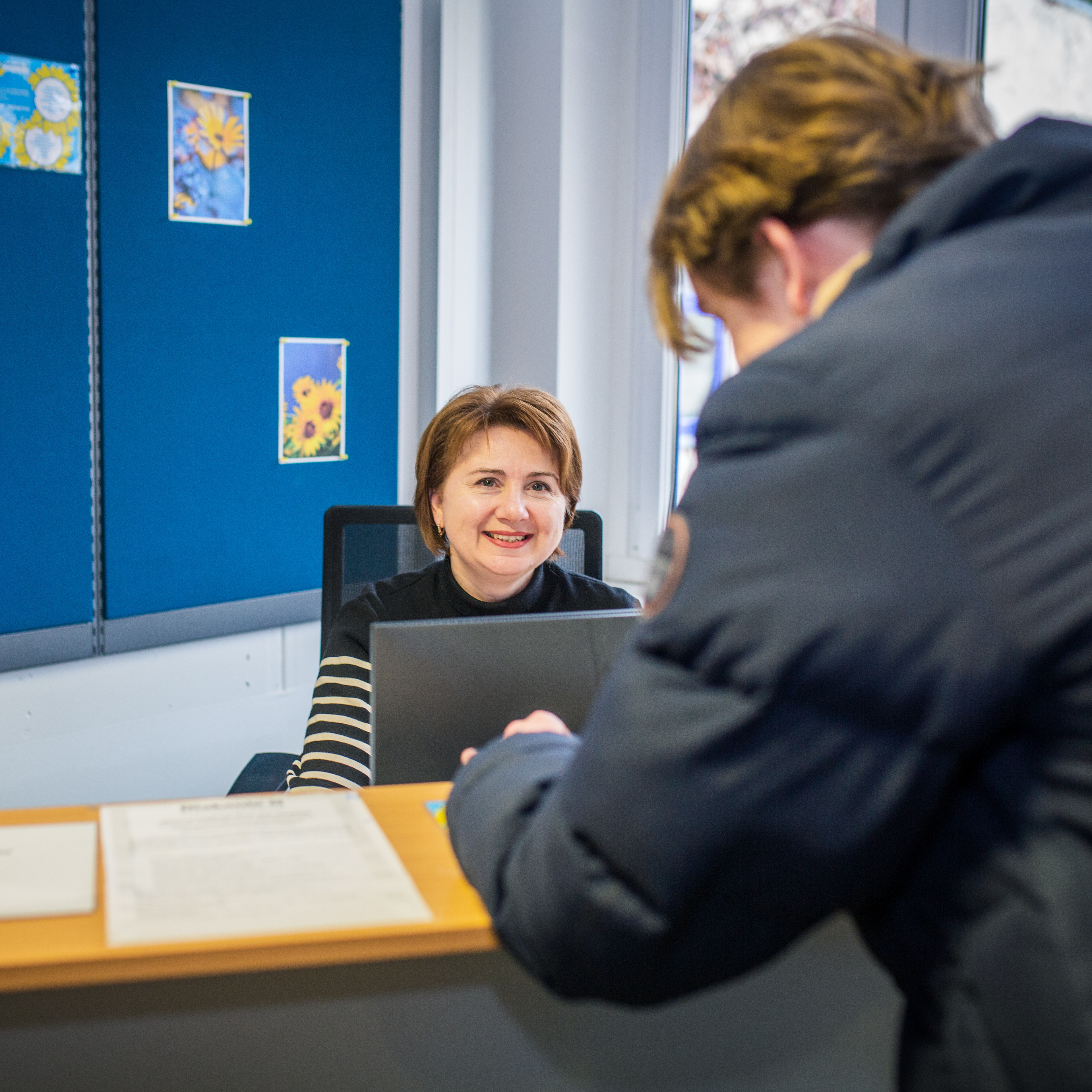 Eine lächelnde Frau sitzt hinter einem Empfangstresen am PC. Man sieht eine weitere Person von hinten am Tresen stehen.