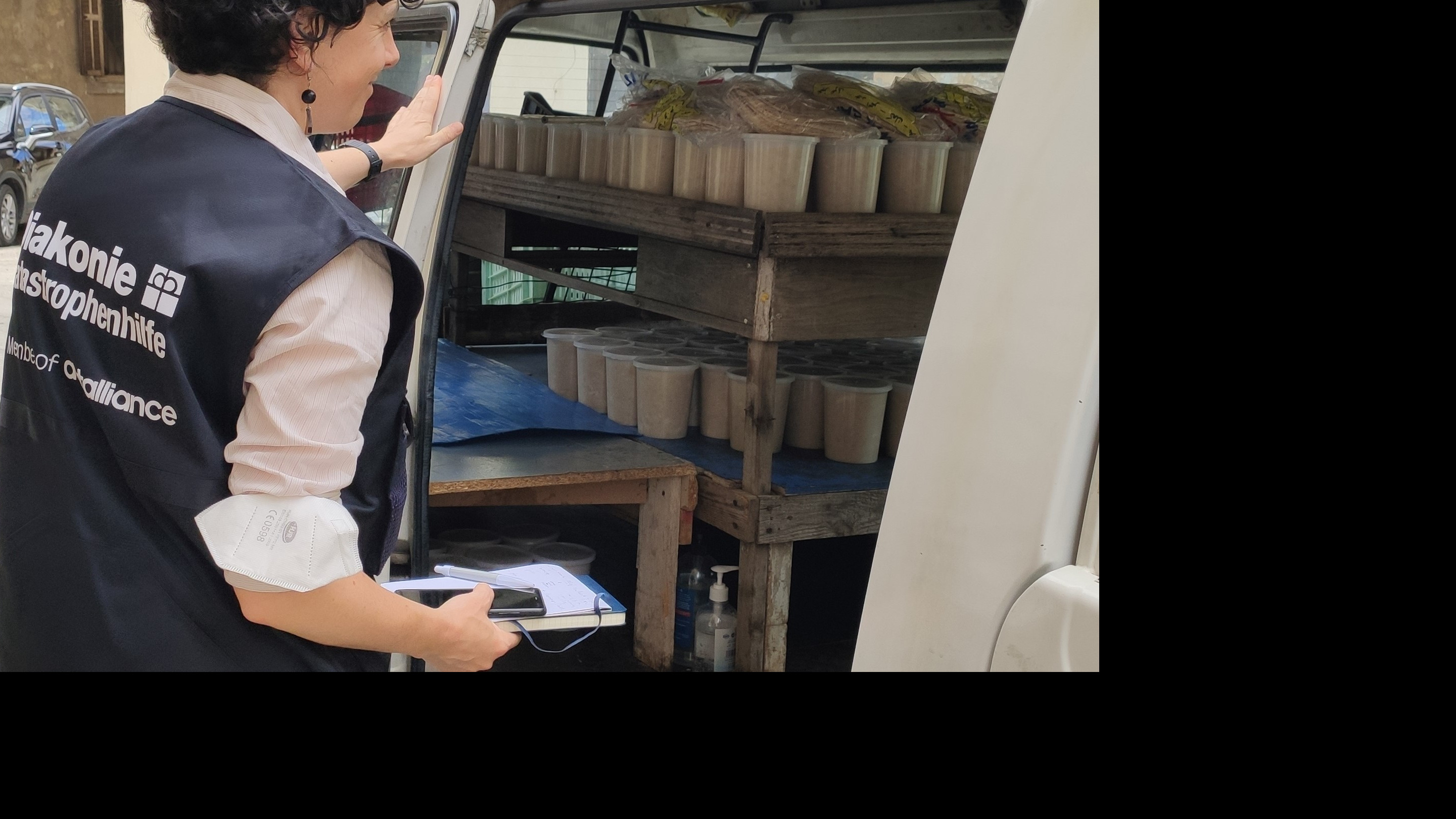 Mitarbeiterin der Diakonie Katastrophenhilfe steht vor einem Auto, das mit portionierten Mahlzeiten und Fladenbrot beladen ist.