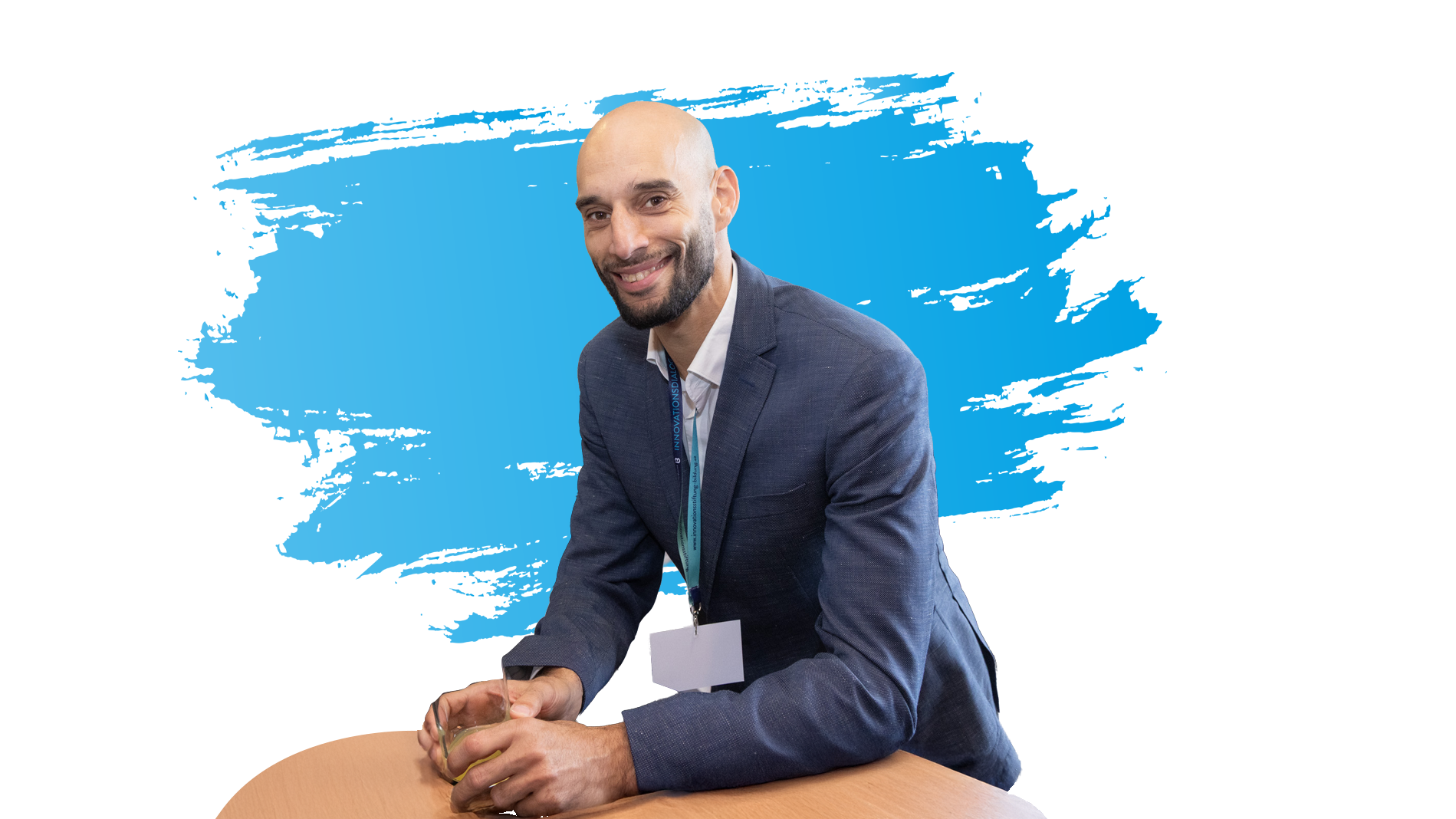 Ein Mann lehnt an einem Tisch und lächelt in die Kamera. Im Hintergrund ist ein blauer Farbstrich.