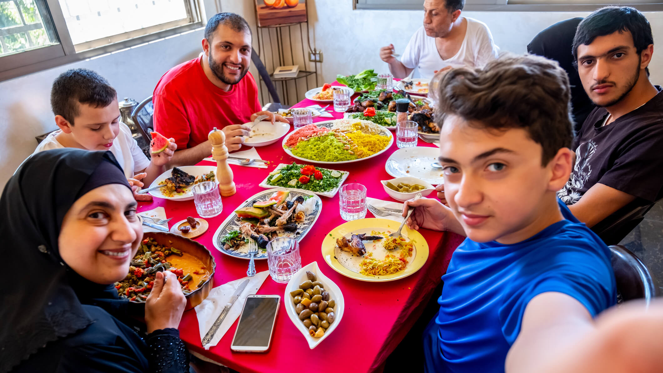 Familie sitzt gemeinsam am Esstisch und isst. Ein junger Mann macht ein Selfie mit der Familie, während alle lächelnd in die Kamera blicken.