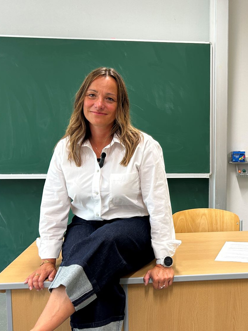 Frau sitzt auf Lehrerpult. Im Hintergrund grüne Schultafel und Skelett.