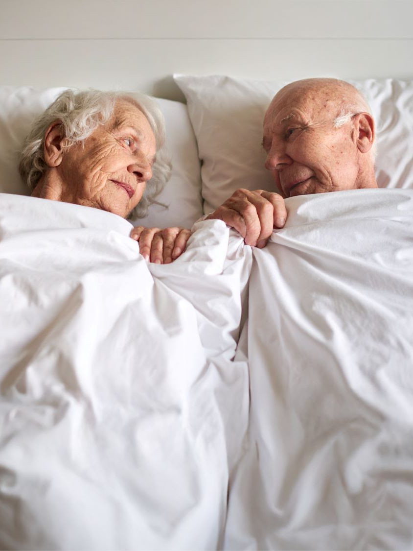 Ein älteres Paar zugedeckt mit weißer Bettdecke blickt sich in die Augen