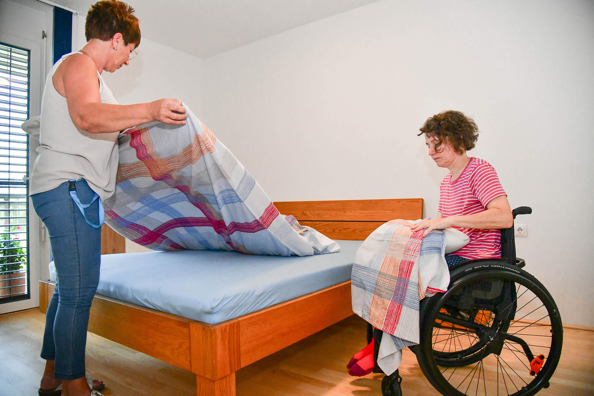 Zwei Frauen machen ein Bett. Eine der Frauen sitzt im Rollstuhl.
