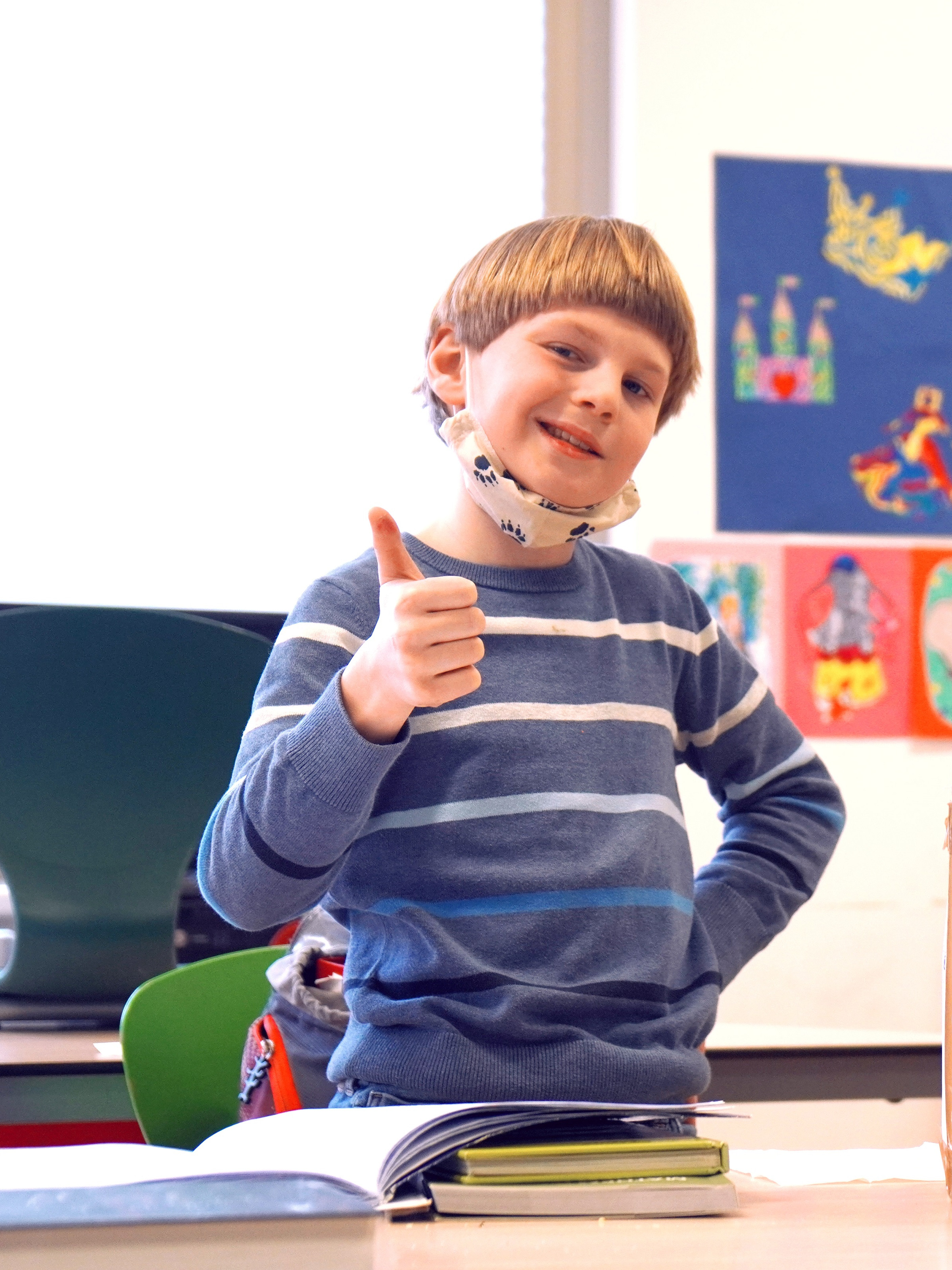 Ein junger Schüler zeigt mit dem Daumen nach oben und schaut stolz in die Kamera, neben ihm steht auf dem Schultisch eine selbstgebastelte Aktentasche aus Karton, auf der in großen Buchstaben Akten steht.