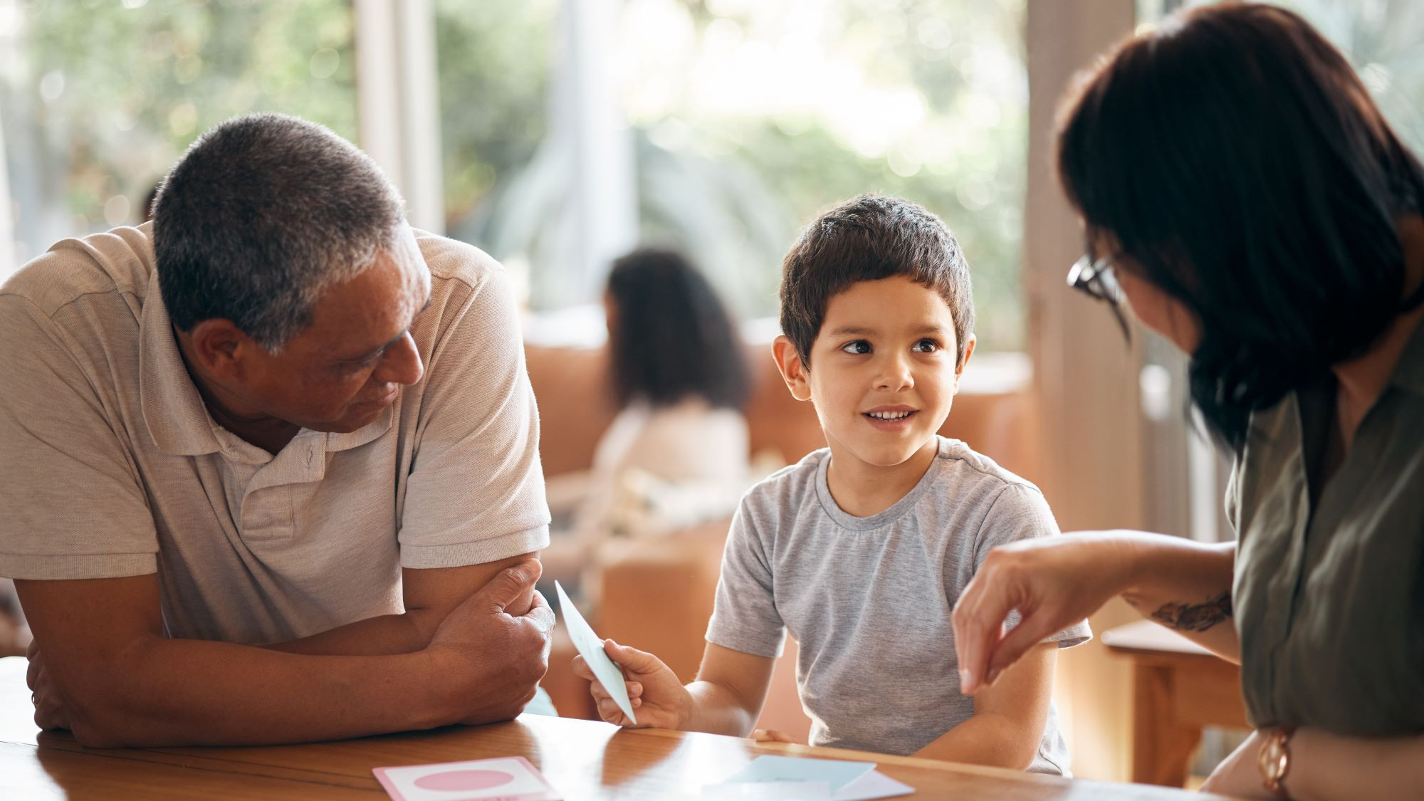 Kleiner Junge sitzt mit zwei Erwachsenen am Tisch und zeigt Kärtchen