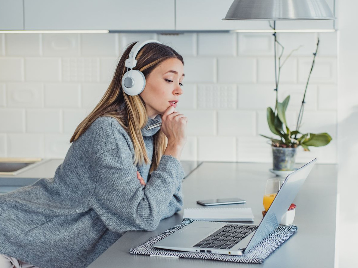 Dame in hellgrauem Pullover und Headset sitzt beim Schreibtisch vor aufgeklapptem Laptop