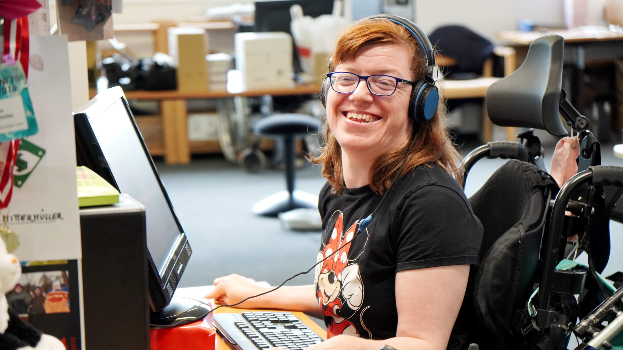 Foto einer Person im Rollstuhl die hinter einem Computer sitzt und in die Kamera lächelt